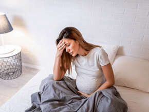 Mujer embarazada sentada en la cama, con aspecto estresado y la mano en la frente.
