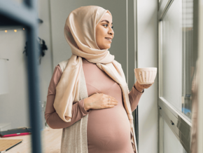 Photograph: pregnant woman in headscarf holding mug, hand on belly, looking out window.