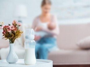 Madre amamantando al bebé en un sofá, con biberón preparado al lado.