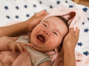 Fotografía de bebé llorando envuelto en toalla rosa, manos adultas sosteniendo su cabeza