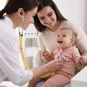 Madre conversando con la pediatra en consulta, ambas inclinadas hacia el bebé. Tono de decisión informada y acompañada, iluminación natural. Sin producto visible, sin logos.