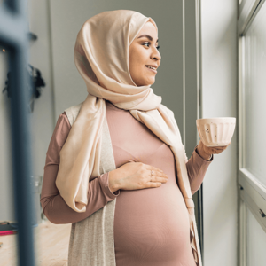 Photograph: pregnant woman in headscarf holding mug, hand on belly, looking out window.