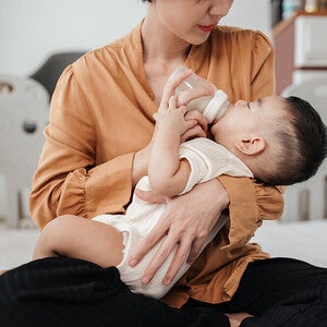 Photograph of mother giving a bottle to an infant on bed, tender expression, crib in background