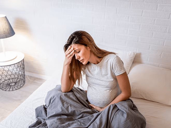 Mujer embarazada sentada en la cama, con aspecto estresado y la mano en la frente.