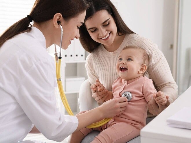 Madre conversando con la pediatra en consulta, ambas inclinadas hacia el bebé. Tono de decisión informada y acompañada, iluminación natural. Sin producto visible, sin logos.