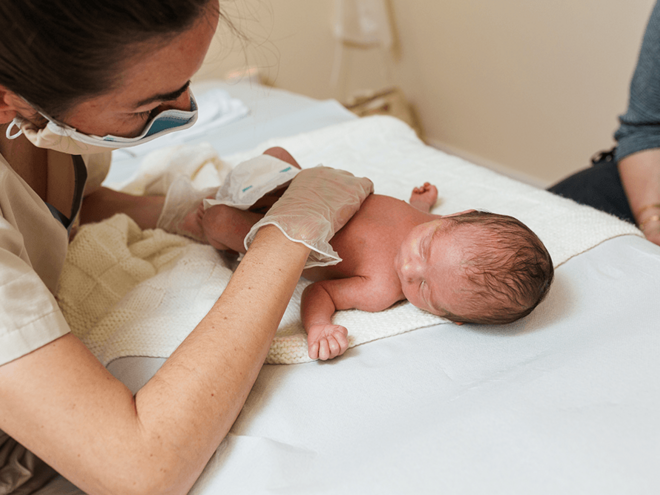 Photo: nurse in gloves examines a newborn lying on a white sheet while parent watches