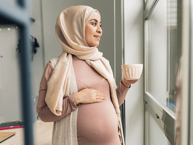 Photograph: pregnant woman in headscarf holding mug, hand on belly, looking out window.