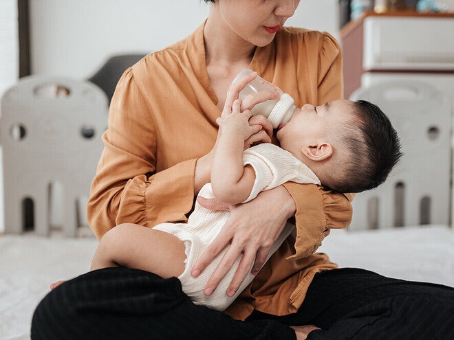 Photograph of mother giving a bottle to an infant on bed, tender expression, crib in background