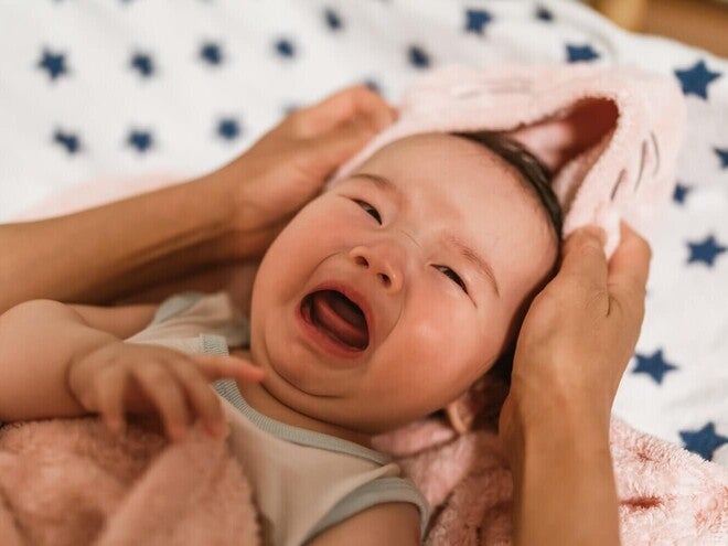 Fotografía de bebé llorando envuelto en toalla rosa, manos adultas sosteniendo su cabeza