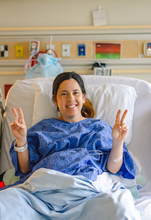 Fotografía de mujer sonriente en cama de hospital, con bata azul, haciendo el signo de paz