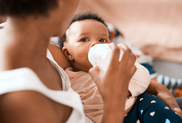 Photo: adult feeding infant with bottle, baby gazing at camera