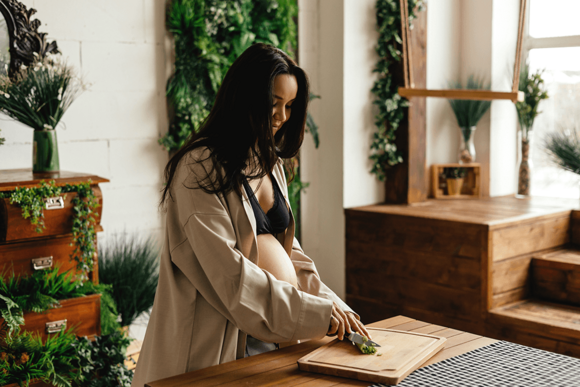 Photograph of a woman chopping herbs on a wooden board in a sunlit, plant-filled kitchen